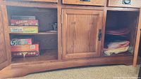 Oak cabinet showing games stacked on the left side lower compartment and folded table linens in middle and right compartments.