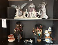 General overview of all figurines and decorative items on two shelves including stained glass angels, Kachina dolls, 'LOVE' sign, toy truck, pottery, and owl figurines.