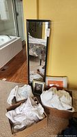 Photo showing full view of tall black framed wall mirror leaning against yellow wall, three boxes of bedding and towels below it, and a framed floral picture on the floor.