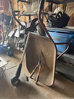 Metal wheelbarrow showing rust on tray and wheel, Black & Decker string trimmer leaning against it, Fiskars shears resting inside wheelbarrow