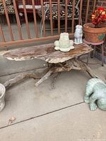 Wood table with live edge top and tree root base shown in patio setting with decorative ceramic frogs and plant pots around.