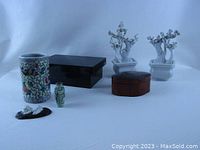 Photo showing all items in lot arranged on a white surface: black lacquer box, floral lacquer trinket box, enamel brush pot, two white Blanc de Chine cherry tree figures, small carved snuff bottle, and porcelain foo dog on wooden base.