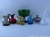 Photo of all nine assorted colored glass pieces arranged in a row including cruets, pitchers, compote, and dresser jar.
