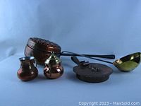 Five assorted copper and brass kitchen items arranged together showing size and proportion, includes popcorn maker with perforated lid, ladle, sugar and creamer, crumb catcher