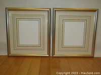 Pair of gold-toned gilt wood frames shown side by side resting on wooden floor, each with intricate multi-layer mats in shades of pink and green and blank interior openings.