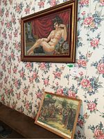 Photo showing both framed art pieces hanging and leaning against floral wallpapered wall; larger needlepoint above, smaller historical print below.