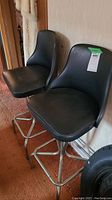 Two black vinyl swivel seat bar stools seen side by side from front angle on carpeted floor, showing seats and backs.
