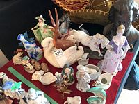 Overview of various ceramic, wood, and resin figurines on a red tablecloth including lady, swan, horse, birds, and busts.
