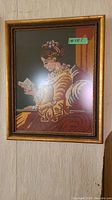 Framed needlepoint of woman reading a letter in a simple gold frame under glass.