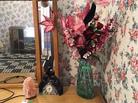 Full view of the lot placed on wooden table with floral wallpaper background showing green glass vase with faux pink florals, ceramic clock, and pink salt lamp.