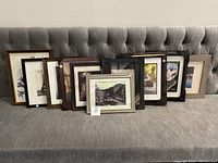 Ten framed artworks grouped on a tufted gray sofa, includes black, brown, and beige frames with various print subjects such as flowers and buildings.