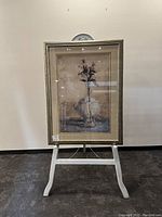 Full view of framed floral art print on white stand against a beige wall and dark floor.