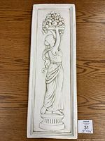 Heavy stone rectangular plaque with raised relief of a woman carrying a fruit basket over her head in classical attire, placed on wooden surface.