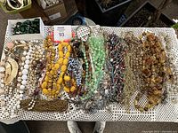 Wide shot of assorted antique necklaces laid out on a crocheted fabric, showing various bead colors, sizes, and textures including yellow, green, white, and amber tones.
