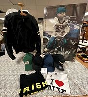 Front view of black leather motorcycle jacket on hanger with scattered caps and t-shirts on table beneath.