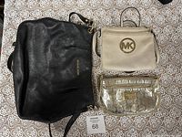 Three Michael Kors purses grouped on lace patterned surface: large black shoulder bag, beige crossbody bag with MK logo, and small metallic gold pouch with embossed logos.