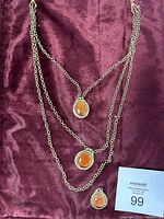 Two silver-tone chain necklaces with amber-like stone pendants, displayed on burgundy fabric, along with a small matching pendant charm and a lot card.