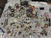Top-down view of a large collection of assorted antique and vintage jewelry laid out on a white lace tablecloth, featuring bracelets, brooches, earrings, and other small pieces.