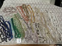 Wide assortment of vintage necklaces laid out on lace fabric including whites, greens, blues, corals, and blacks.