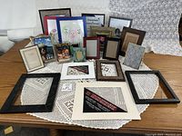 Full view of 19 assorted picture frames arranged on a table with a lace cloth background, showing variety in size, shape, color, and materials.