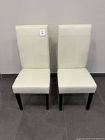 Front view of matching pair of white leather dining chairs with dark wooden legs on tiled floor against white wall.