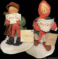 Photo of both Annalee Caroling Couple dolls standing on snow-covered bases, holding Christmas music sheets, with visible tags.