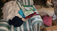 Various vintage clothing items laid on a striped sofa, showing knitted, crocheted, and fabric garments in cream, pink, and blue colors.