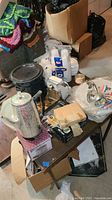 Photo of vintage and miscellaneous kitchenware on a table in basement setting including metal meat grinder, plastic cups, napkins, coasters, and various boxed items