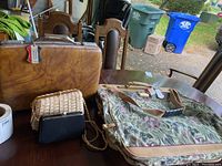 Image showing the full lot including the tapestry garment bag, attaché briefcase, wicker purse, and black evening bag on wood table.