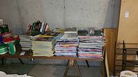 Large collection of magazines stacked on a wooden table in a basement area with concrete walls.