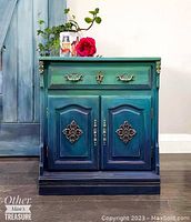 Front view of the boho style cabinet showing green and blue gradient paint, drawer with brass handles and two doors with metal decorative accents.