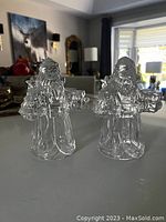 Two glass Santa Claus shaped candle holders standing side by side on a reflective surface with a background of home interior.