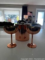 Copper-colored metal ice bucket with lid shown alongside two matching copper-colored metal champagne glasses on a white surface with living room background.