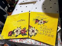 Two square canvas paintings laid side by side on a polka dot tablecloth. Both feature bright yellow chalk paint backgrounds, prominent floral designs in red, white, and green, and decorative scripted text.