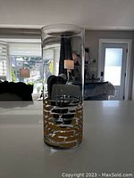 Tall clear glass cylindrical vase sitting on a white surface, showing gold horizontal stripes design near the bottom.