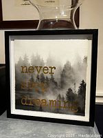 Photo of framed print with black square frame and artwork showing misty forest and gold lettering.