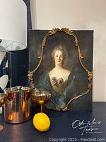 Front view of the canvas art showing a portrait of a woman with an ornate wood molding frame and dark background, placed next to copper and silver drinkware and a lemon.