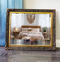 Full front view of large square ornate mirror resting on floor, showing carved dark bronze frame with gold inner edge and reflective glass.