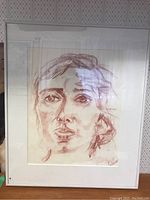 Full view of the framed signed portrait drawing showing the person's face with expressive brown/red sketch lines, white matting, metal frame, and glass with reflections.