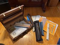 Photo showing open brown briefcase containing music books, recorders in cases/pouches, and metal stand on wood floor.