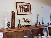 Wide view of all figurines and bronze items on wooden shelf, including camel, geese, fisherman, bell, white candle holder, and wooden carved piece.