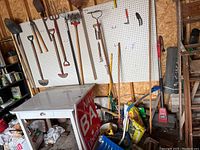 Full view of garden tools hanging and leaning against pegboard and wall including shovels, rakes, pitchfork, spade, broom, with bags on floor and a small table
