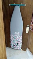Metal ironing board with blue frame, grey fabric cover with pink and white floral bottom design hung on door.