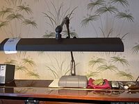 Full frontal view of the lamp showing the ribbed horizontal shade, weighted base, and adjustable arm on a wooden surface with background wallpaper.