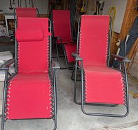 Four red outdoor loungers with black metal frames and armrests. Loungers have red fabric seating with laced sides attached by grommets. One shows visible wear and damage. Four matching red cushions are included.