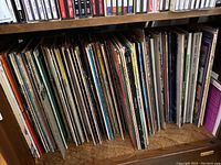 Shelf with over 40 vinyl LP records showing a variety of album covers and edges stored upright on wood shelf.