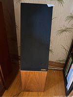 Front view of one speaker with grille installed showing wood veneer lower section and black grille cover.