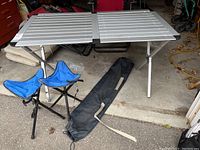 Collapsible aluminum metal table with two blue foldable stools and a black carrying bag placed on a concrete floor near garage entrance.
