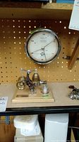 Toy steam engine model on a wooden base with metal and brass parts, next to a round pressure gauge on pegboard.