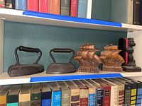 Photo of two cast iron irons with wooden handles, wooden ship-shaped bookends, and wooden beaver shoe brush holder on a shelf next to books.